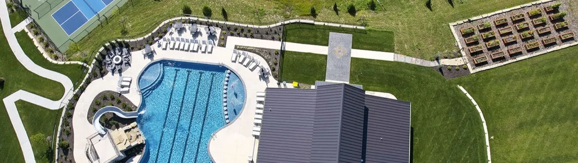An overhead aerial view of the Century Farms community in Overland Park, Kansas: a freeform swimming pool with a slide, a dark-roofed clubhouse, pickleball courts, a community garden, walking paths and lawns, in summer