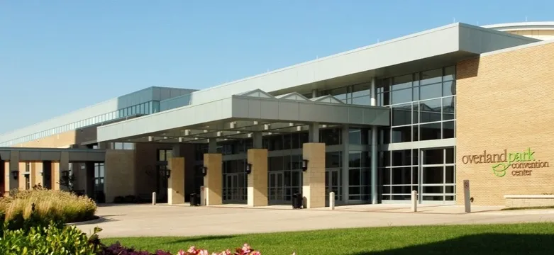 The Overland Park Convention Center in Overland Park, Kansas, a long tan brick building with a glass entrance canopy, a landscaped flower bed and a green lawn under a clear sky