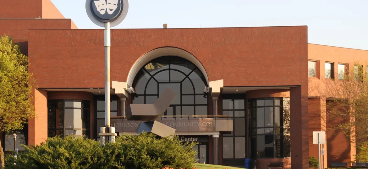 Johnson County Community College, Overland Park, Kansas: the Carlsen Center's brick entrance with an arched window, a round theater-mask sign on a pole, and a sculpture in front of the college's name lettering