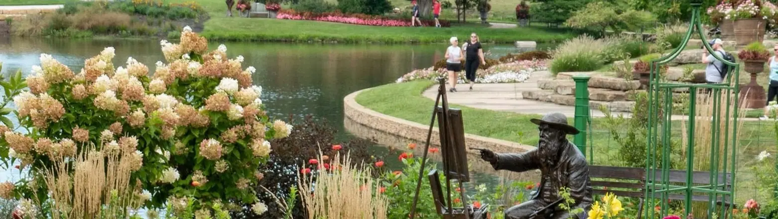 Overland Park Arboretum and Botanical Gardens, Overland Park, Kansas: a bronze statue of a painter at an easel among flower beds beside a pond, with the visitor center beyond