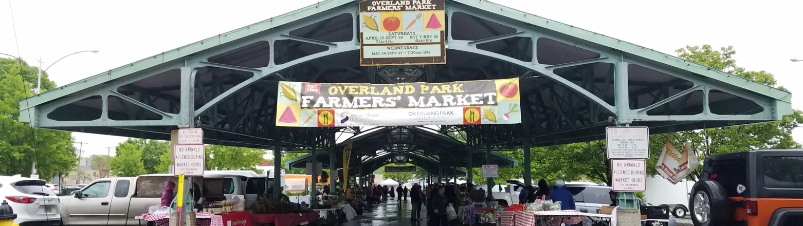 Overland Park Farmers' Market, Overland Park, Kansas: the covered market pavilion with vendor tables and the market's banner, on a rainy day