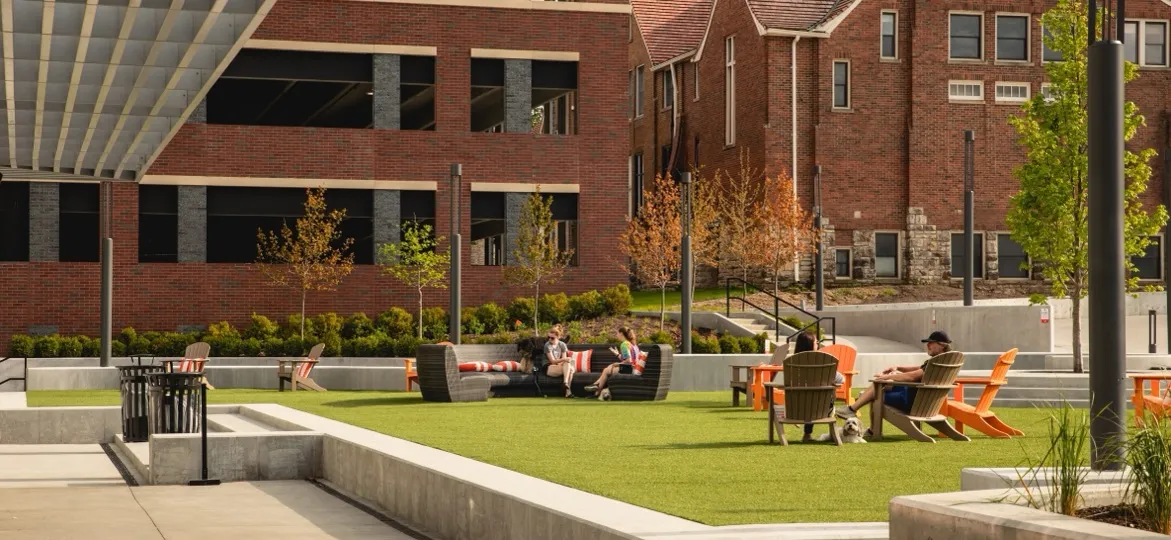A landscaped plaza with a green lawn, lounge seating and orange chairs beside a brick building in Overland Park, Kansas, with people relaxing on the grass