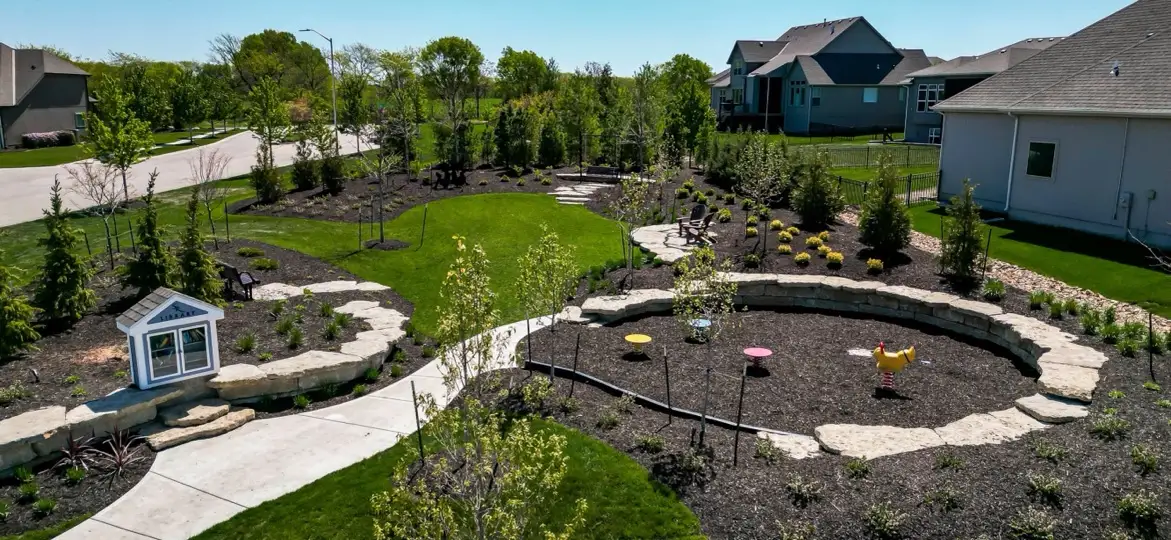 A landscaped neighborhood common area in Overland Park, Kansas, with a winding sidewalk, a stone-walled garden bed, a small playhouse and new homes behind, under a clear blue sky