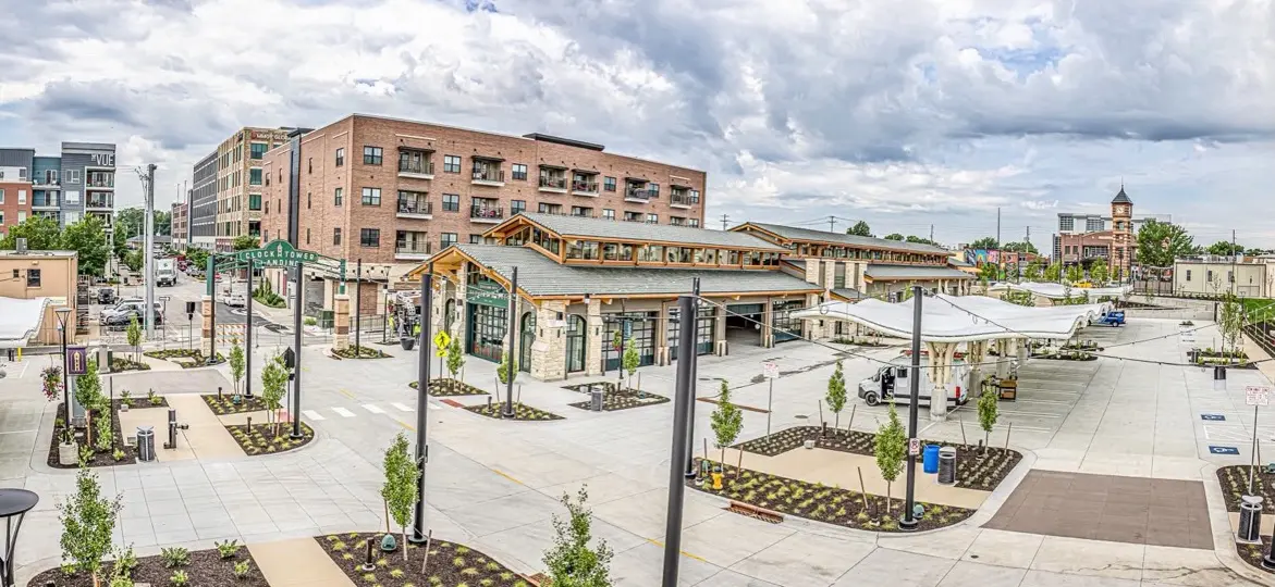 Overland Park Farmers' Market pavilion and open plaza in downtown Overland Park, Kansas, with a brick apartment building and the clock tower in the distance under a cloudy sky