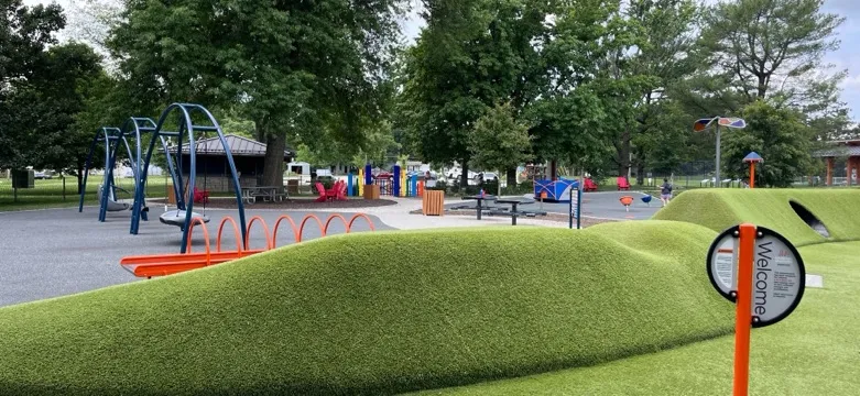 Strang Park in Overland Park, Kansas: a smooth green turf play hill beside a playground with blue swings, an orange seesaw and colorful panels, under mature trees