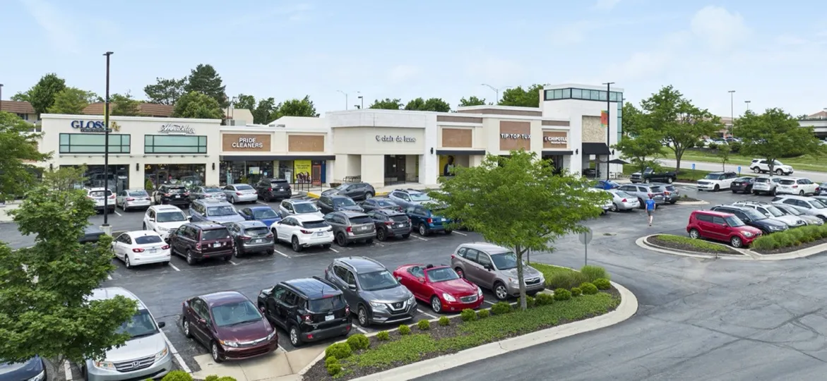 A low shopping center with a row of storefronts, a parking lot and mature trees in Overland Park, Kansas, seen from above on a summer day