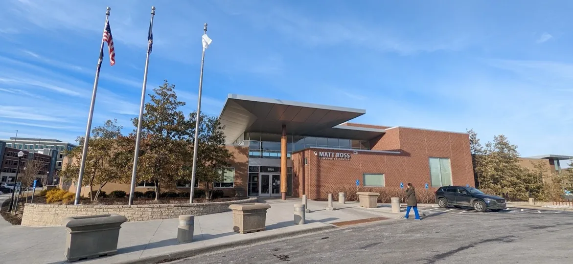 The brick Matt Ross Community Center in Overland Park, Kansas, with a wide metal canopy, three flagpoles and a parking area on a clear blue-sky day