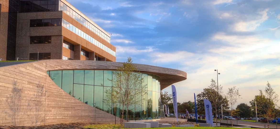 A curved glass and wood-clad entrance pavilion beside a brick office building on the Black & Veatch campus in Overland Park, Kansas, with a lawn in the foreground at dusk